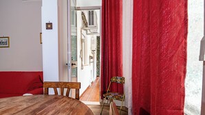 A room with a wooden dining table, a red sofa, a patterned chair, and a window with red curtains.