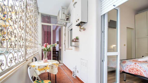 A narrow balcony with a table and chair, a sliding glass door to a bedroom, and a decorative metal fence.