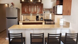 A kitchen with wooden cabinets, a stainless steel refrigerator, a gas stove, a coffee maker, and a blender. There are three bar stools in front of the counter.