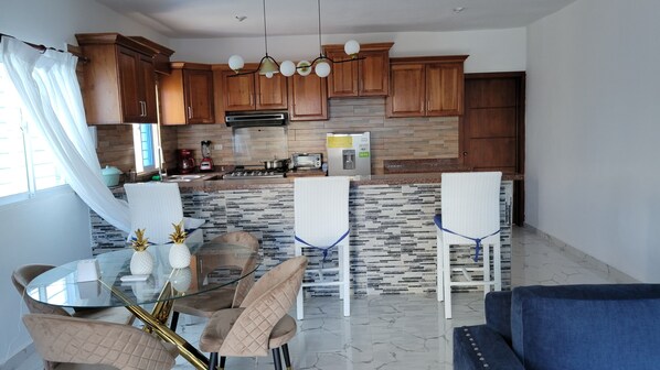 A modern kitchen with wooden cabinets, a built-in oven, and a water dispenser. There is a dining area with a glass table and chairs, and a blue sofa in the living area. - Aparta Hotel Madome (Pedernales)