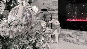 A modern living room with a stone-textured fireplace, a Christmas tree with snow-like decorations, and a white dog statue. - Mountain View Modern Cottage in the Smokies W/ Hot Tub, Newly Renovated. (Sevierville)