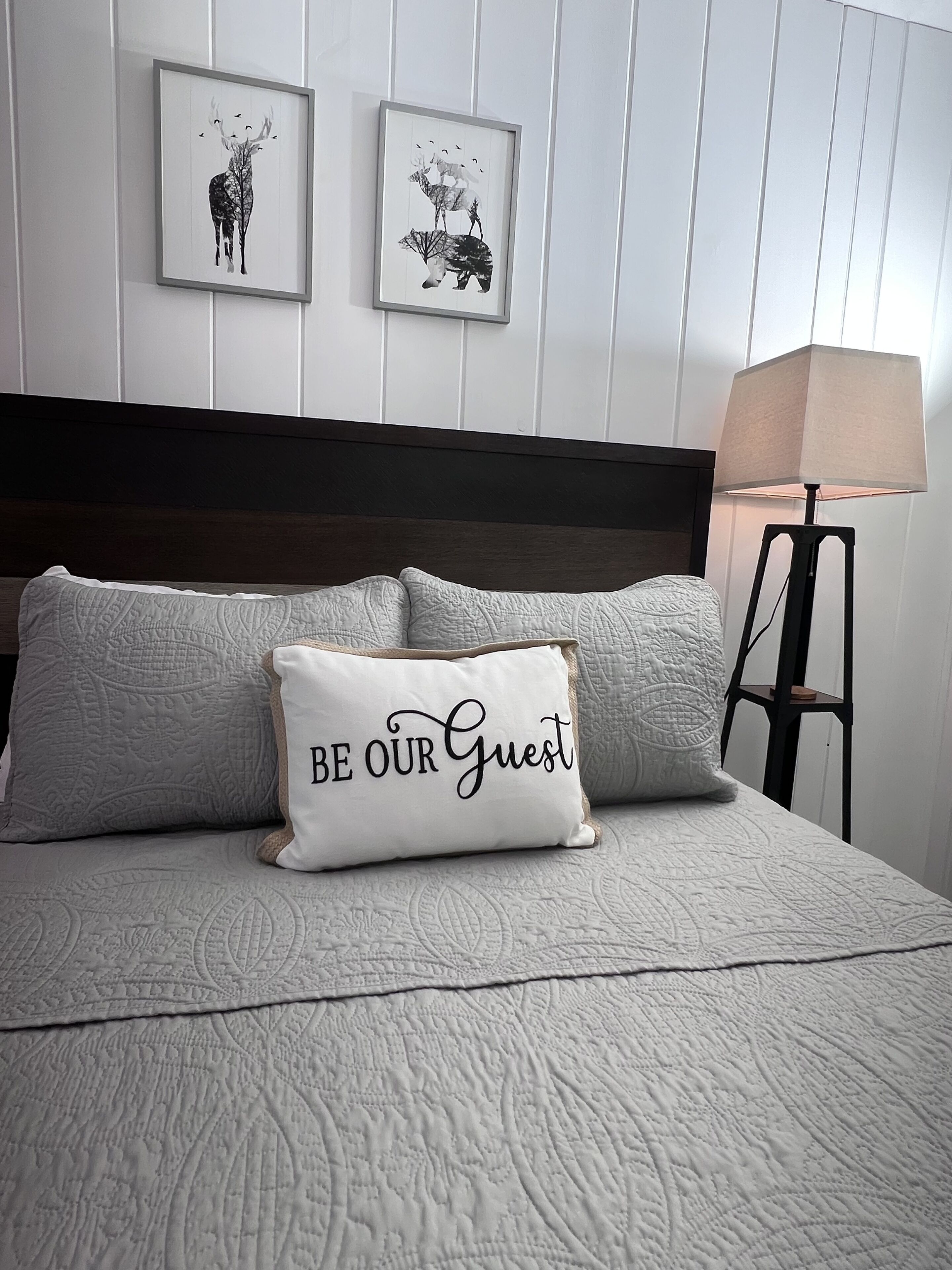 Una cama bien hecha con cubrecama acolchado, dos almohadas y una almohada decorativa con texto. Hay dos cuadros enmarcados en la pared sobre la cama.