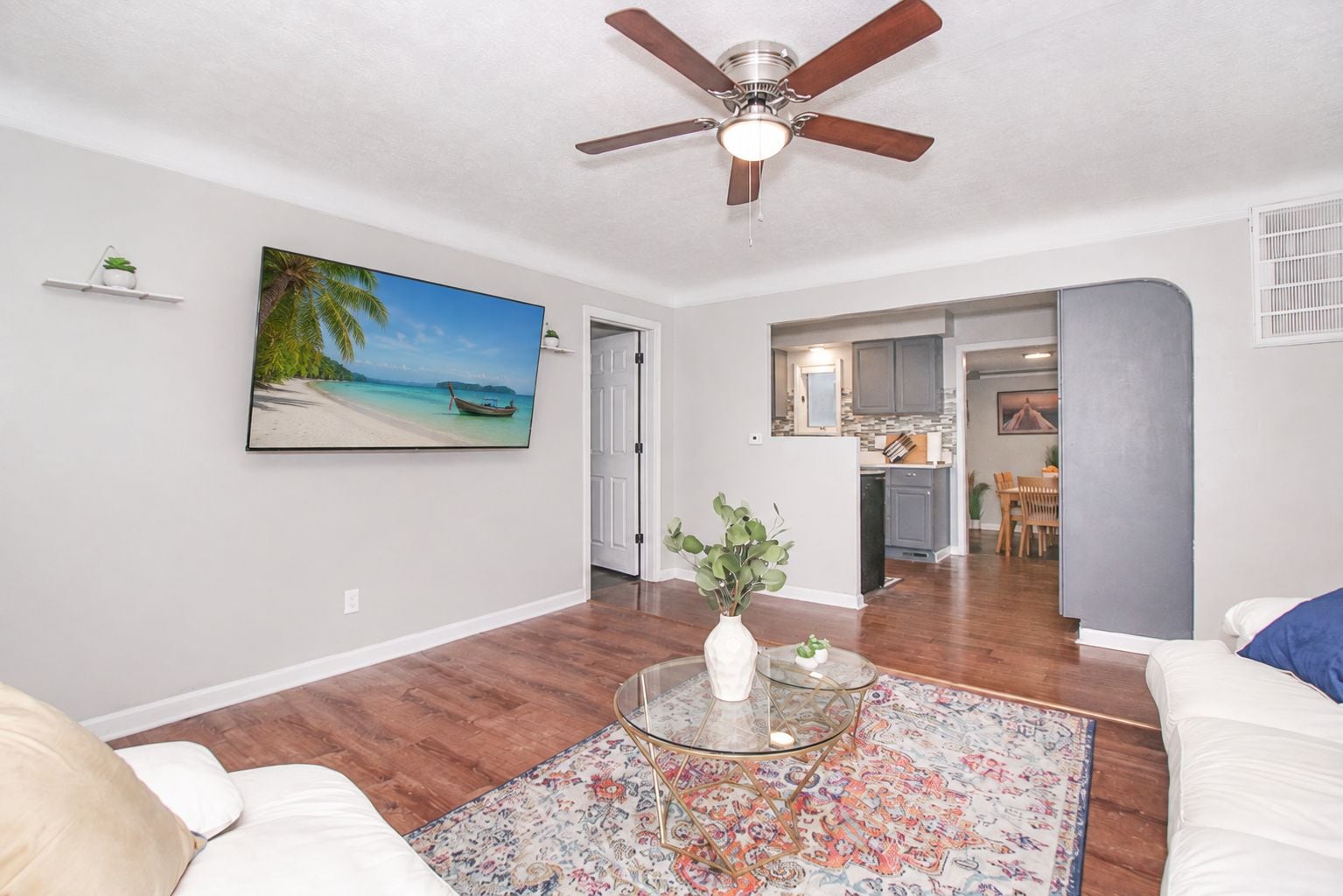 Ein Wohnzimmer mit Deckenventilator, einem Strandbild, einem Essbereich mit Tisch und Stühlen sowie einer Küche im Hintergrund.