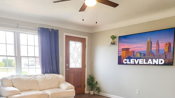 A living room with a beige sofa, a glass coffee table, and a colorful rug. There is a door with a window, a ceiling fan, and a painting of a city skyline. - NICE New Kitchen, King Bed, Fenced Yard, 2 baths, Close to Airport & Downtown (Cleveland)