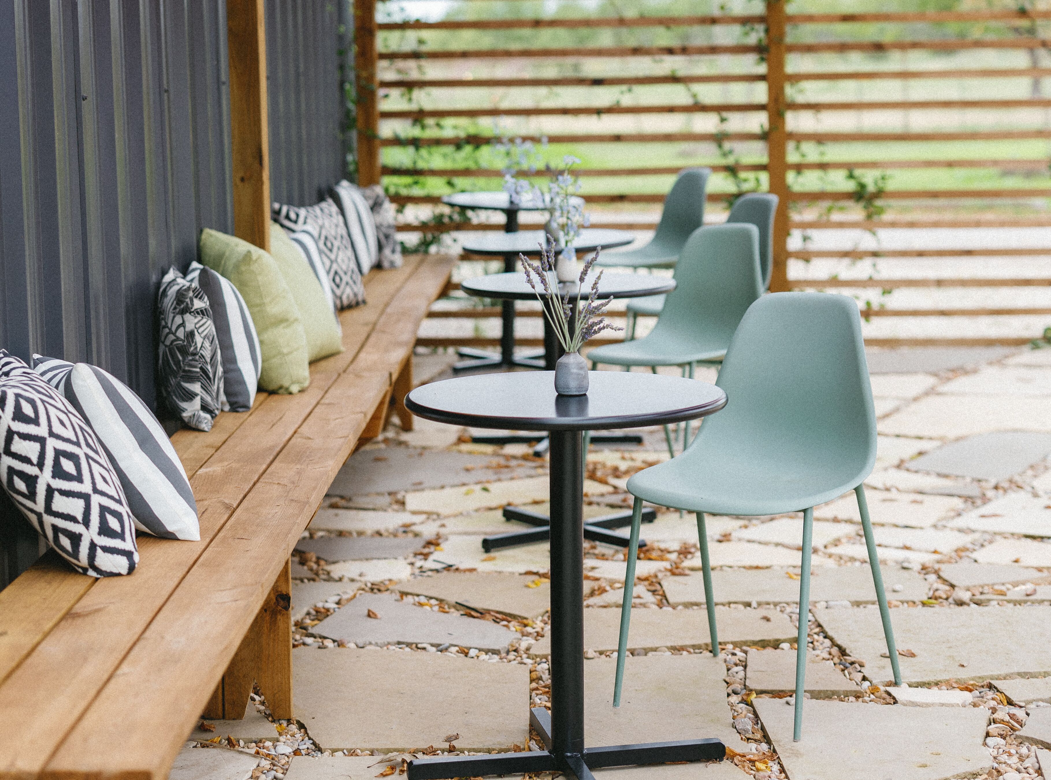 Een overdekt terras met houten banken en ronde tafels, voorzien van gestreepte kussens.