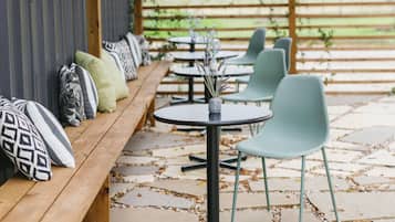 Een overdekt terras met houten banken en ronde tafels, voorzien van gestreepte kussens.