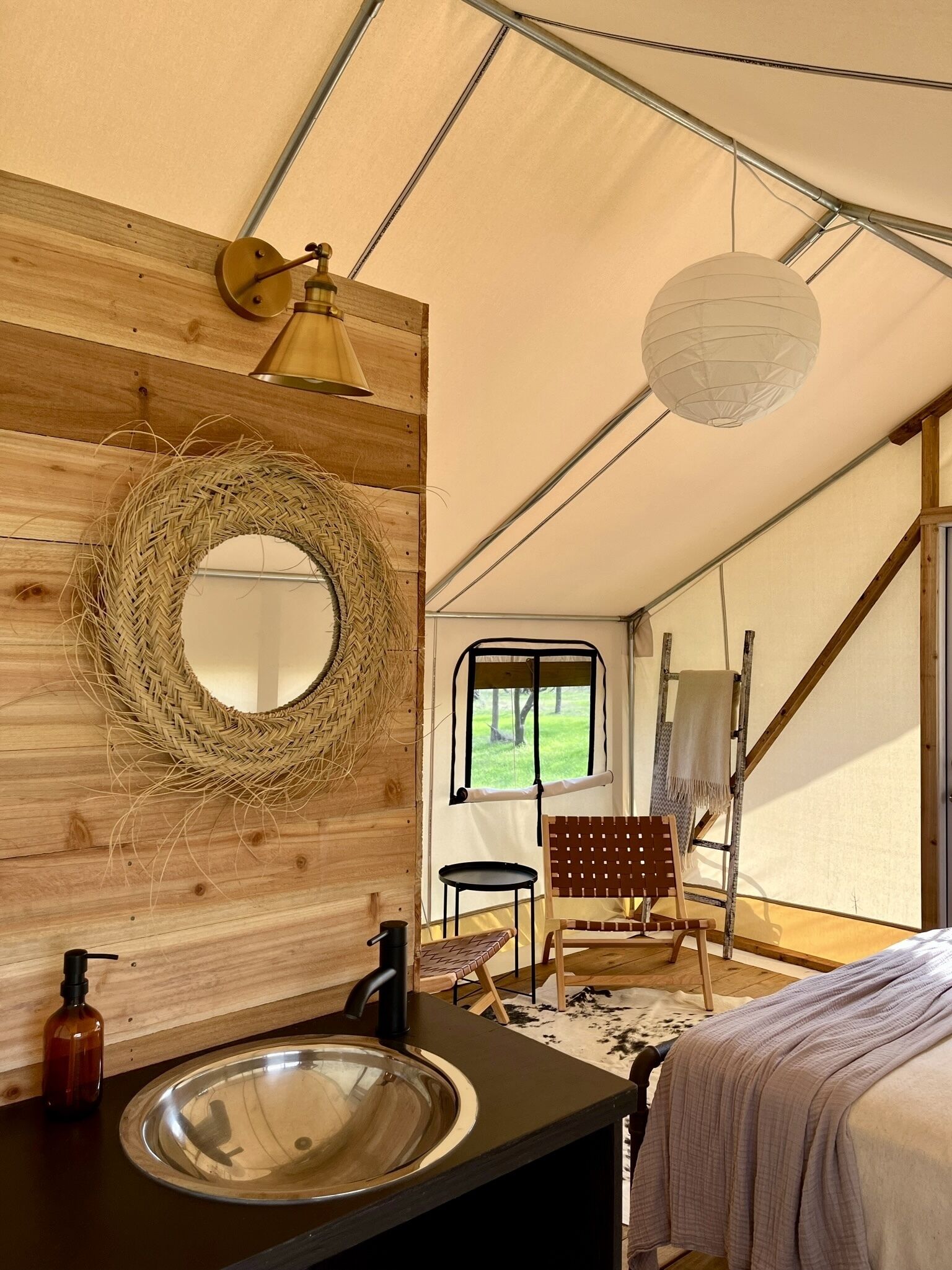 Una carpa con pared de madera, un espejo redondo, un lavabo, una silla, una lámpara y vistas al exterior.