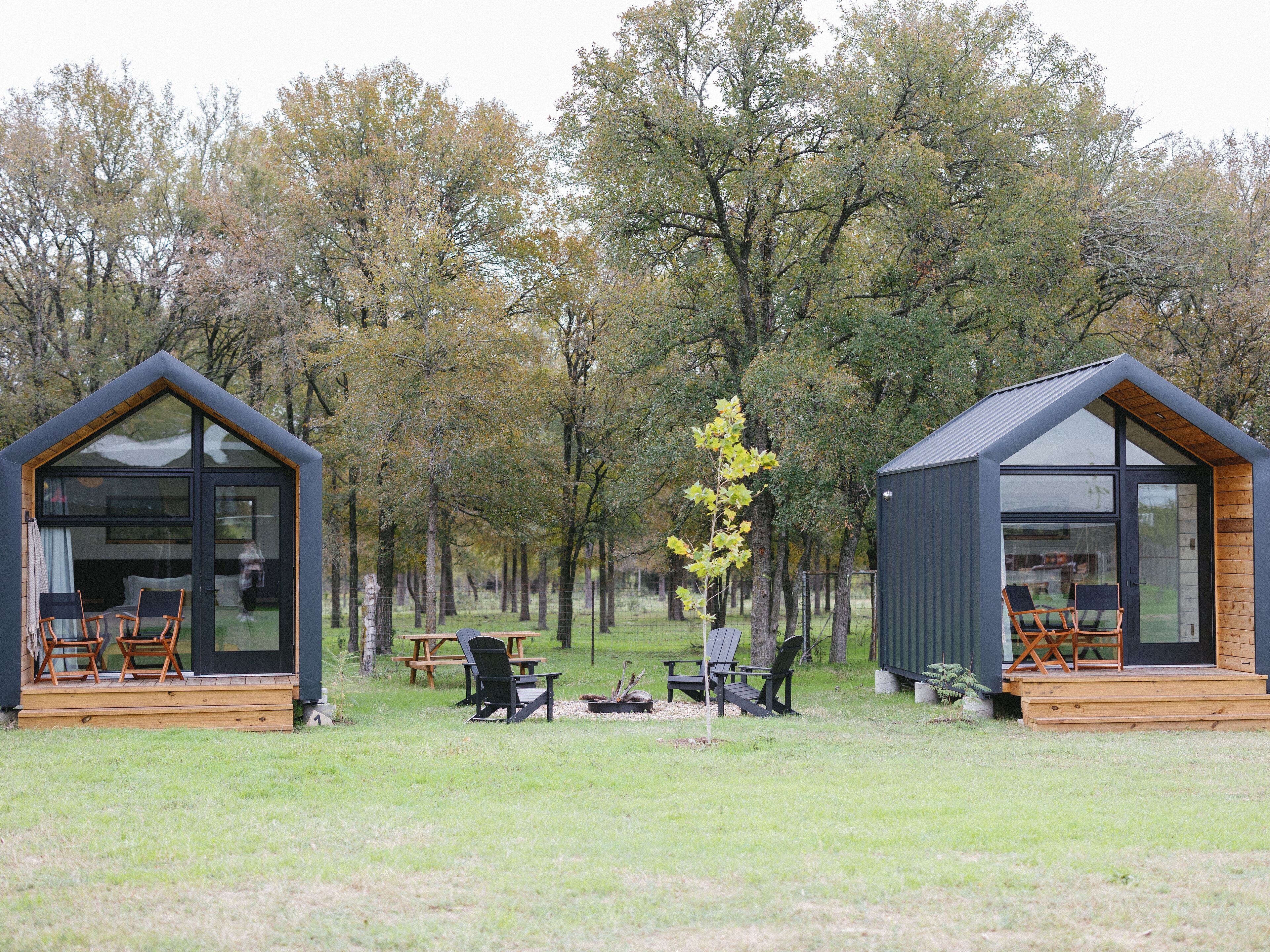 Two small cabins with large windows and wooden decks, surrounded by trees and a picnic table.