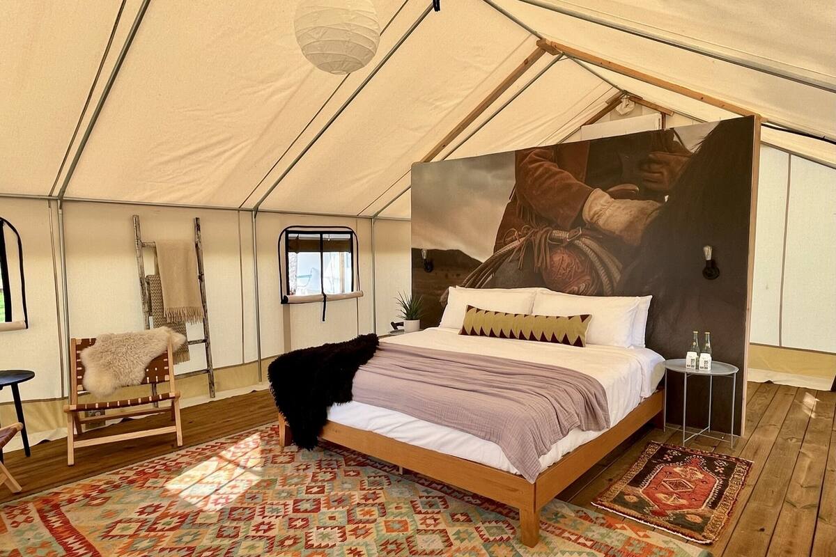 A tented room with a bed, a chair, a small table, and a large tapestry.