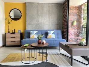 Uma sala de estar moderna com um sofá azul, duas mesinhas laterais, um aparador de madeira e um espelho grande.