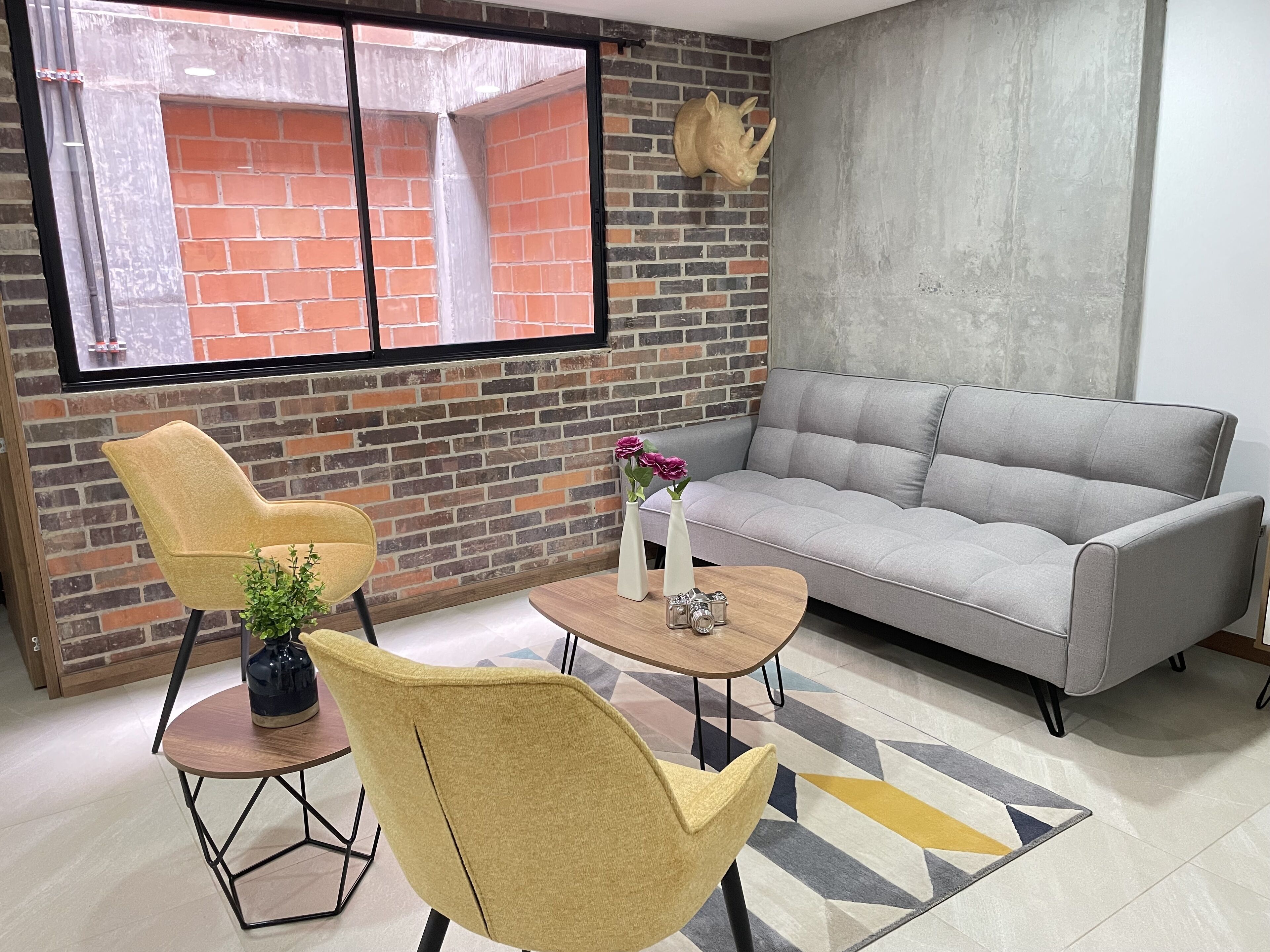 A living room with a brick wall, a grey sofa, yellow chairs, a coffee table with a vase and a camera, and a window with a view of a brick building.