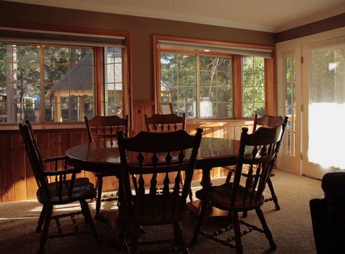 A dining area with a round table and six chairs, large windows with blinds, and a view of trees and a building outside.