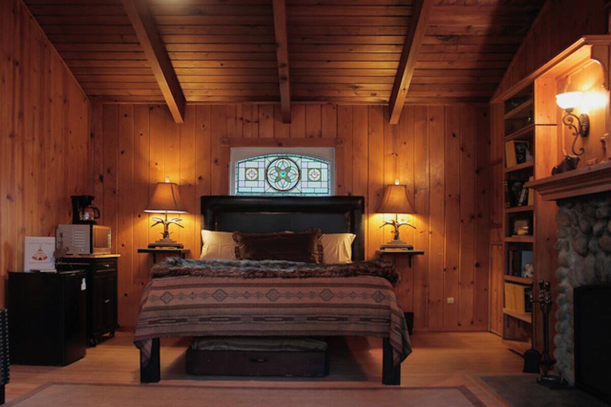 A cozy wooden room with a bed, two bedside lamps, a fireplace, and a stained glass window above the bed.