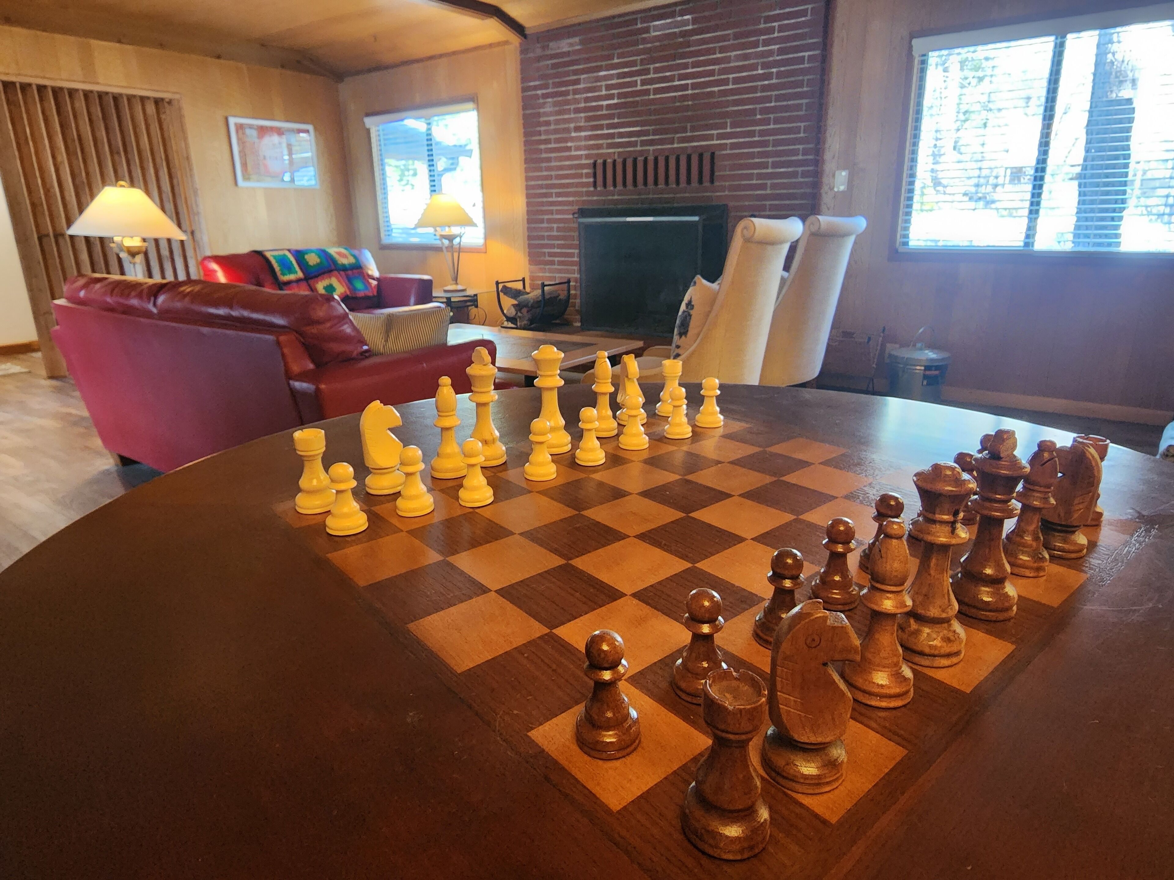 A chessboard with pieces on a wooden table in a room with a brick wall, a fireplace, and a red sofa.