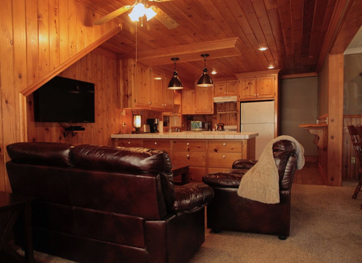 A cozy cabin interior with a wooden ceiling, a fireplace, leather sofas, and a kitchen area.