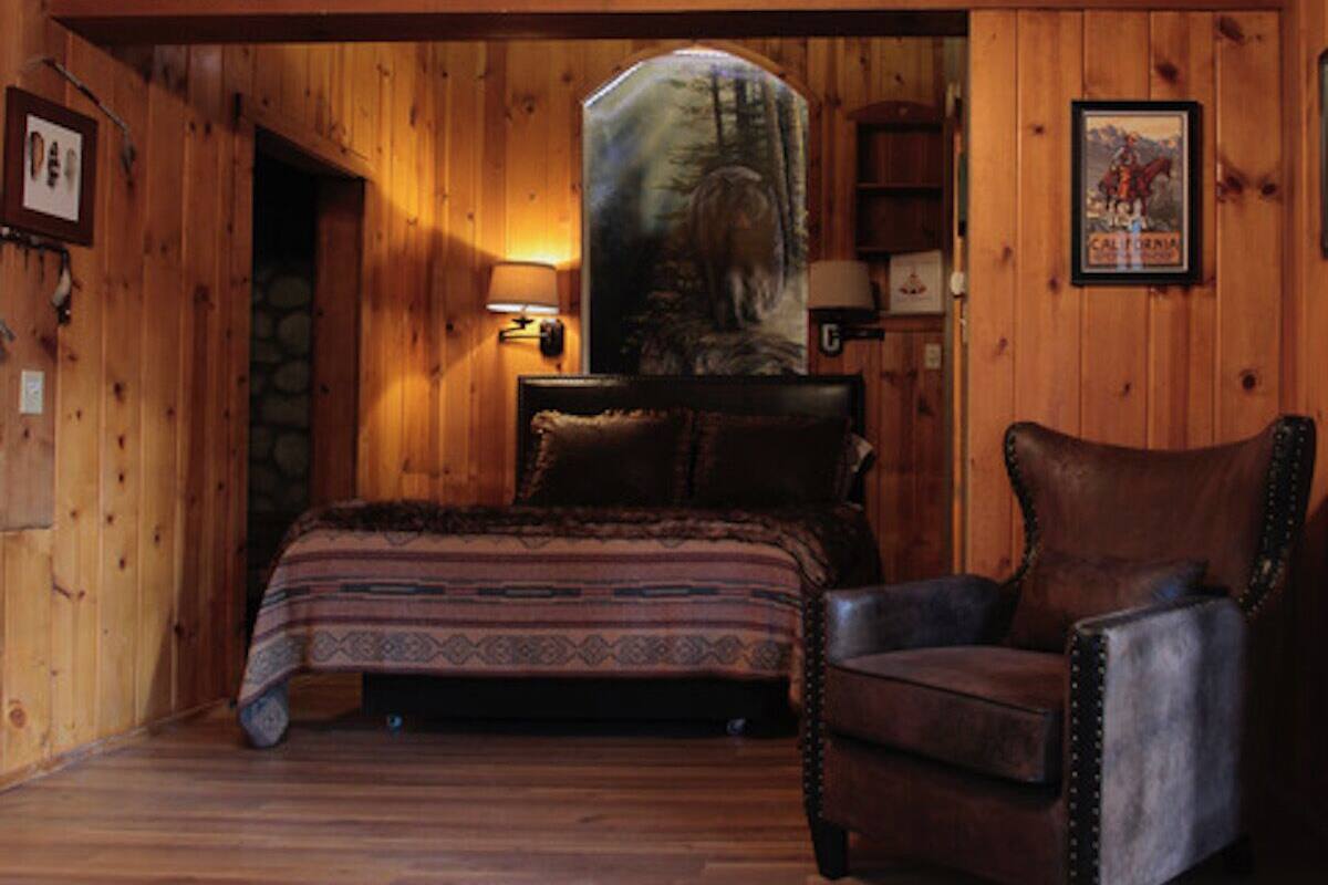 A log cabin room with a bed, a chair, and a painting of a forest scene.