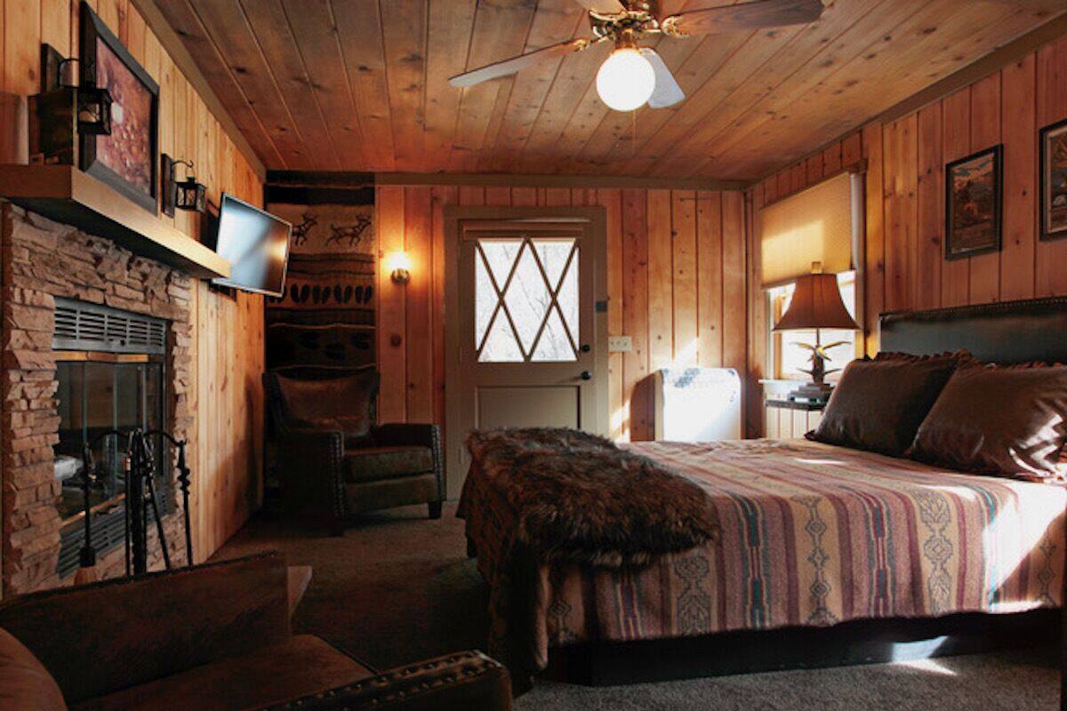 A cozy log cabin bedroom with a stone fireplace, a bed with a patterned blanket, a chair, and a ceiling fan.