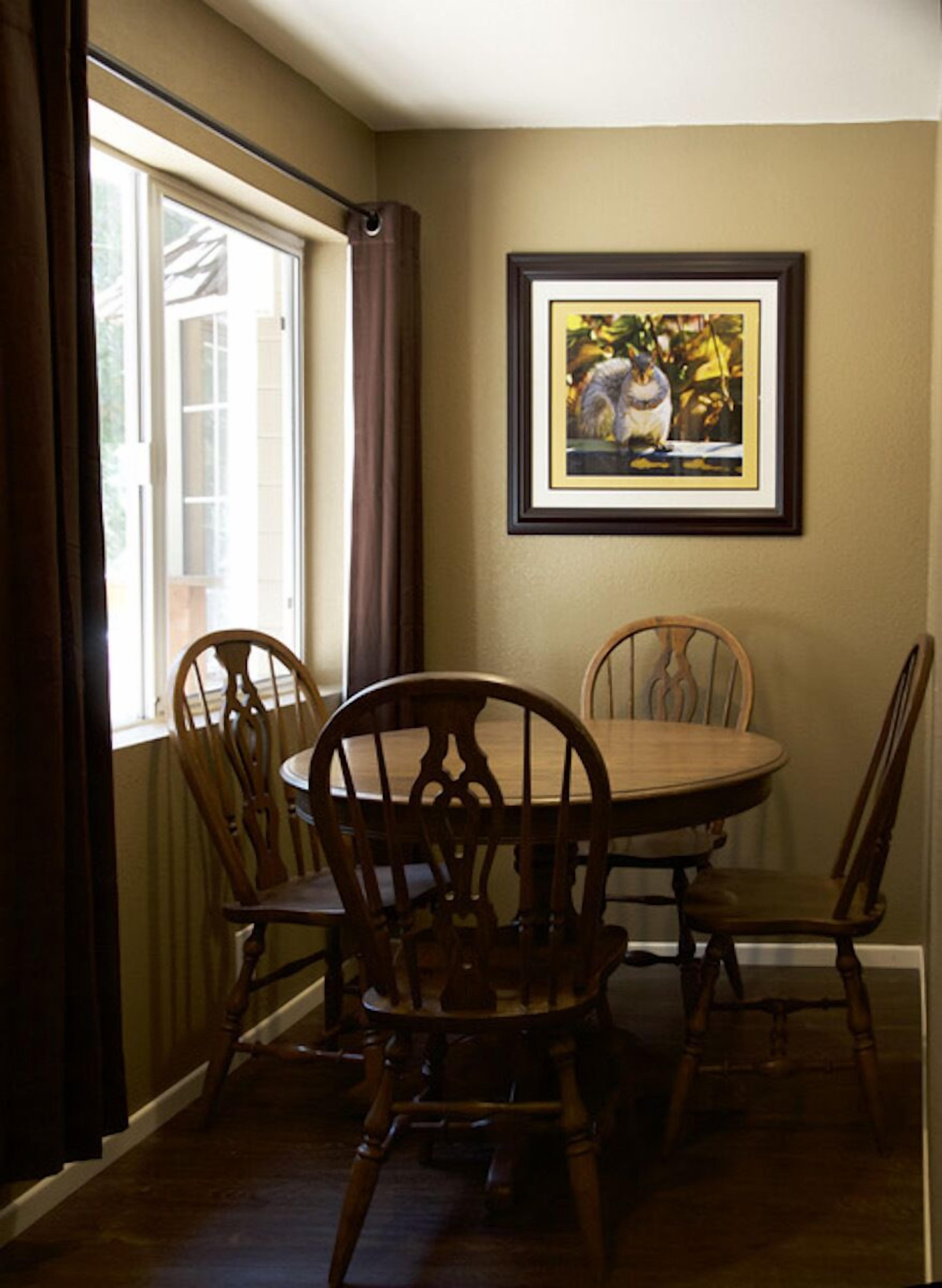A dining area with a round table and four wooden chairs. There is a framed picture on the wall above the table.