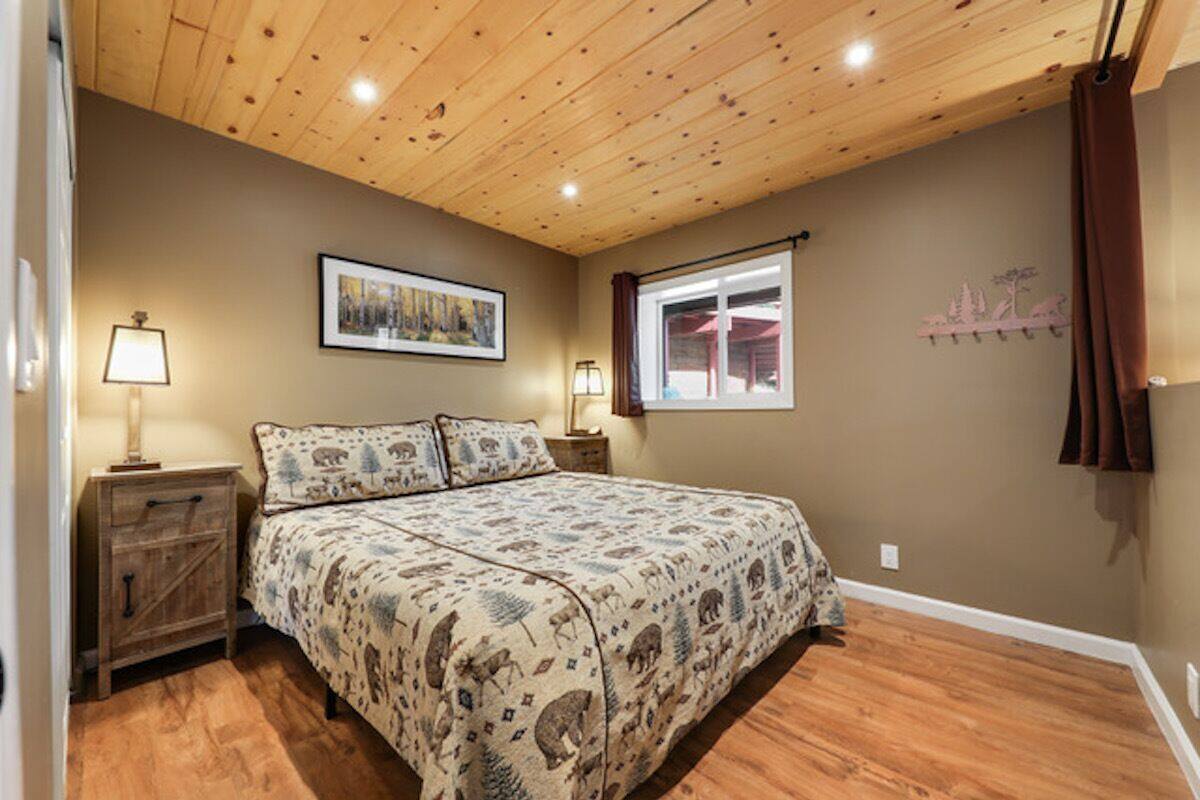 A bedroom with a wooden ceiling, a bed with patterned bedding, a nightstand with a lamp, a window with a view of the outside, and a wall-mounted coat rack.
