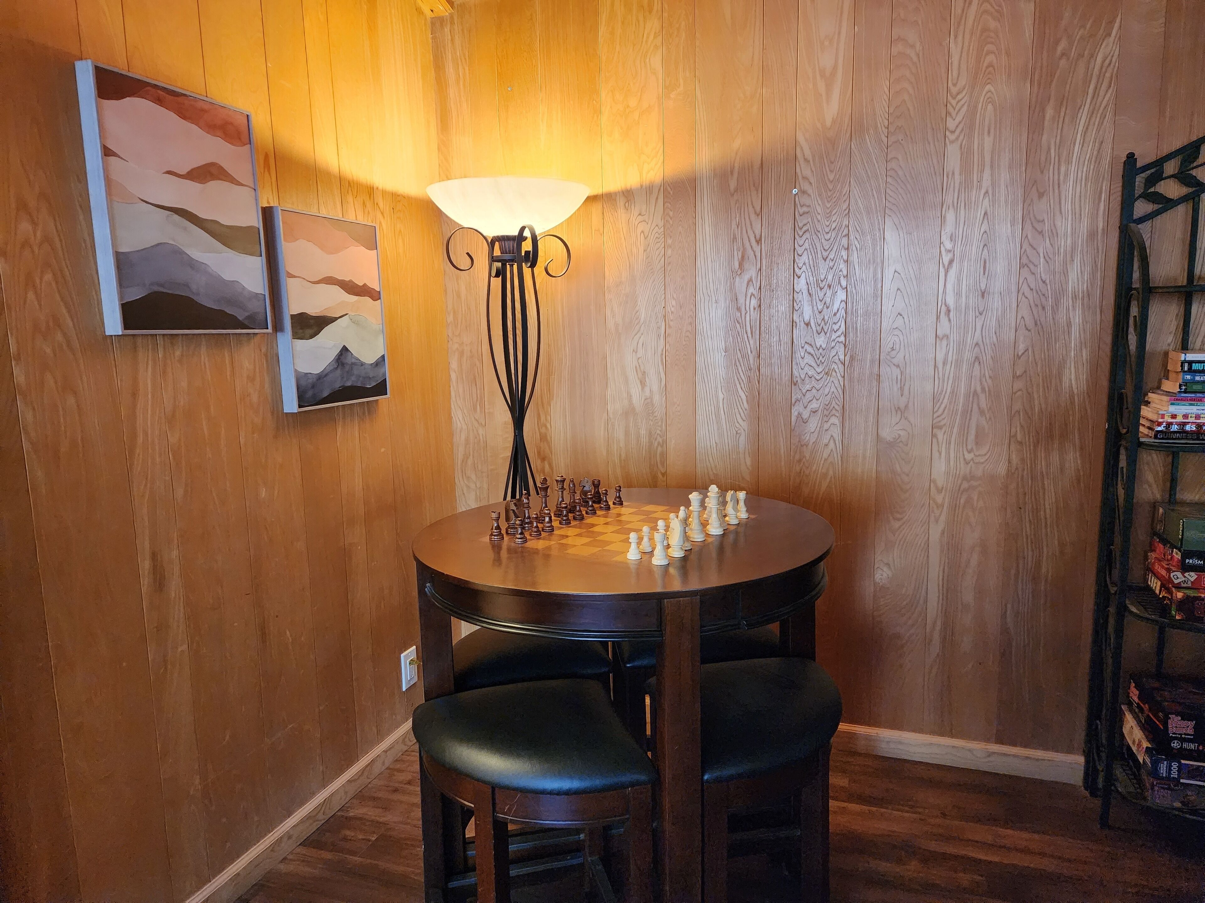 Una habitación con paneles de madera, una mesa redonda dispuesta para un juego, dos sillas, una lámpara y una estantería llena de juegos de mesa.
