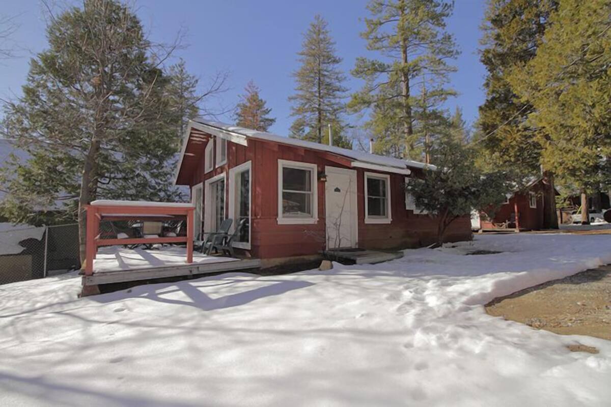 A small, single-story cabin with a red exterior and white trim, surrounded by snow and trees.