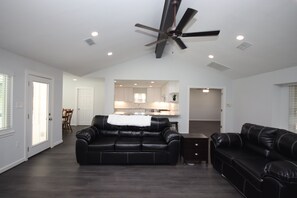 A spacious living room with a black leather sofa, a ceiling fan, recessed lighting, and a view into the kitchen area.