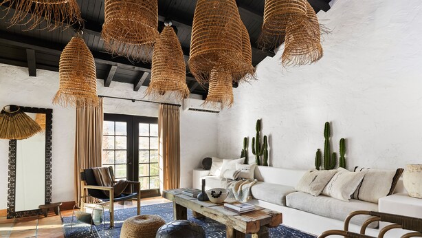 A modern living room with a white sofa, a wooden coffee table, and a blue patterned rug. There are hanging wicker lamps, a large mirror, and several cacti on the wall.