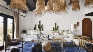 A room with a white sofa, a wooden coffee table, and a blue patterned rug. There are wicker hanging lamps, a cactus wall decoration, and a wooden door.