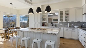 A modern kitchen with white cabinetry, a large island with bar stools, pendant lighting, and a view of mountains through large windows.