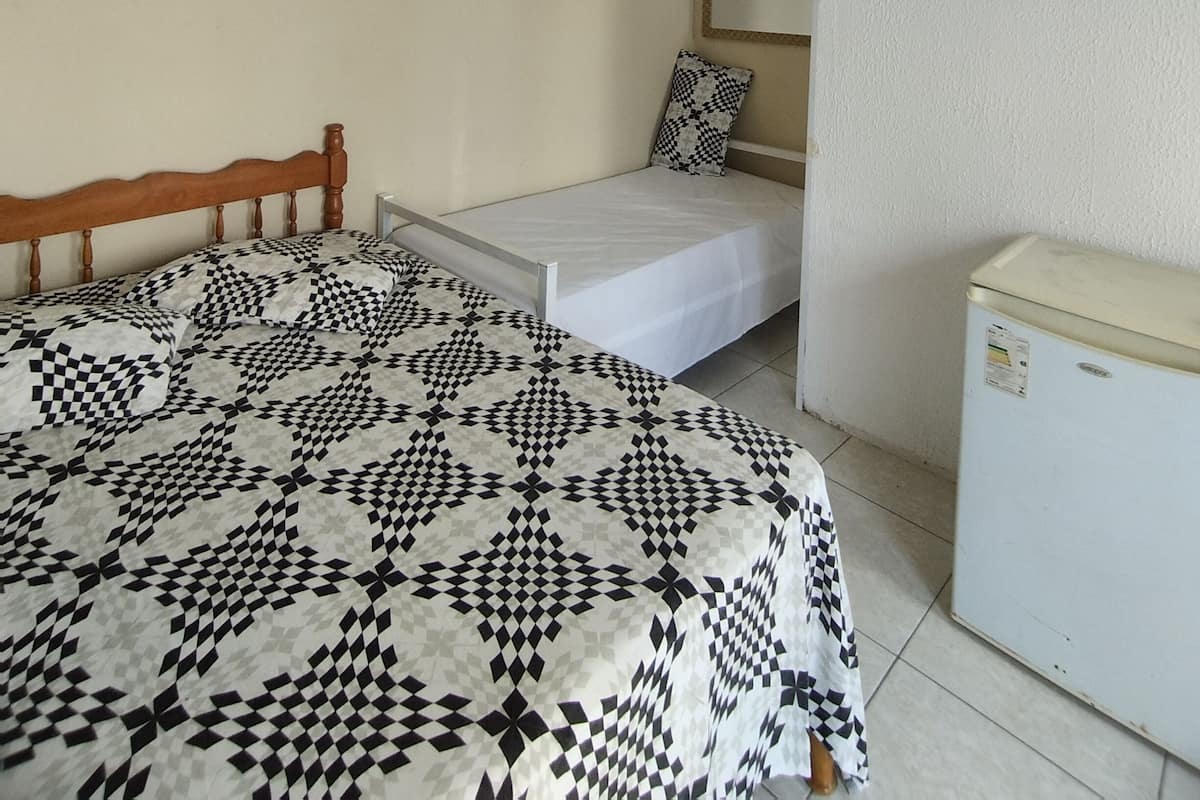 Una habitación pequeña y sencilla con cama, mesita de noche, espejo y refrigerador.