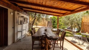 Une pergola en bois installée à l’extérieur, avec une table et des chaises, entourée d’un jardin et d’un bâtiment blanc.
