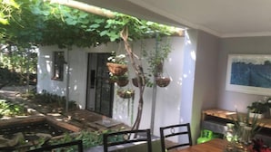 A dining area with a wooden table, chairs, hanging planters, and a view of the garden outside. - Rouana Guest Farm (Stellenbosch)