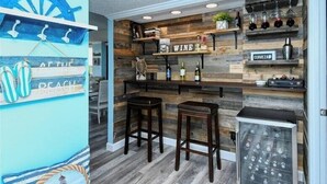 A bar area with wooden shelves displaying bottles and glasses, two stools, and a sign that says "WINE" and "CERVEZA". - Anchorage 404 (Siesta Key)