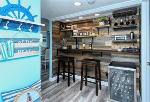 A bar area with wooden shelves displaying bottles and glasses, two stools, and a sign that says "WINE" and "CERVEZA".