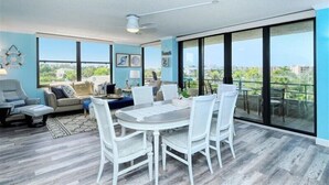 A spacious living area with a dining table, white chairs, a sofa, and a large sliding glass door leading to a balcony. - Anchorage 404 (Siesta Key)