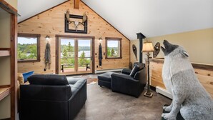 Una habitación acogedora con paredes de madera, techo inclinado y amplios ventanales que ofrecen vistas al exterior. Hay dos sofás de cuero negro y una lámpara.