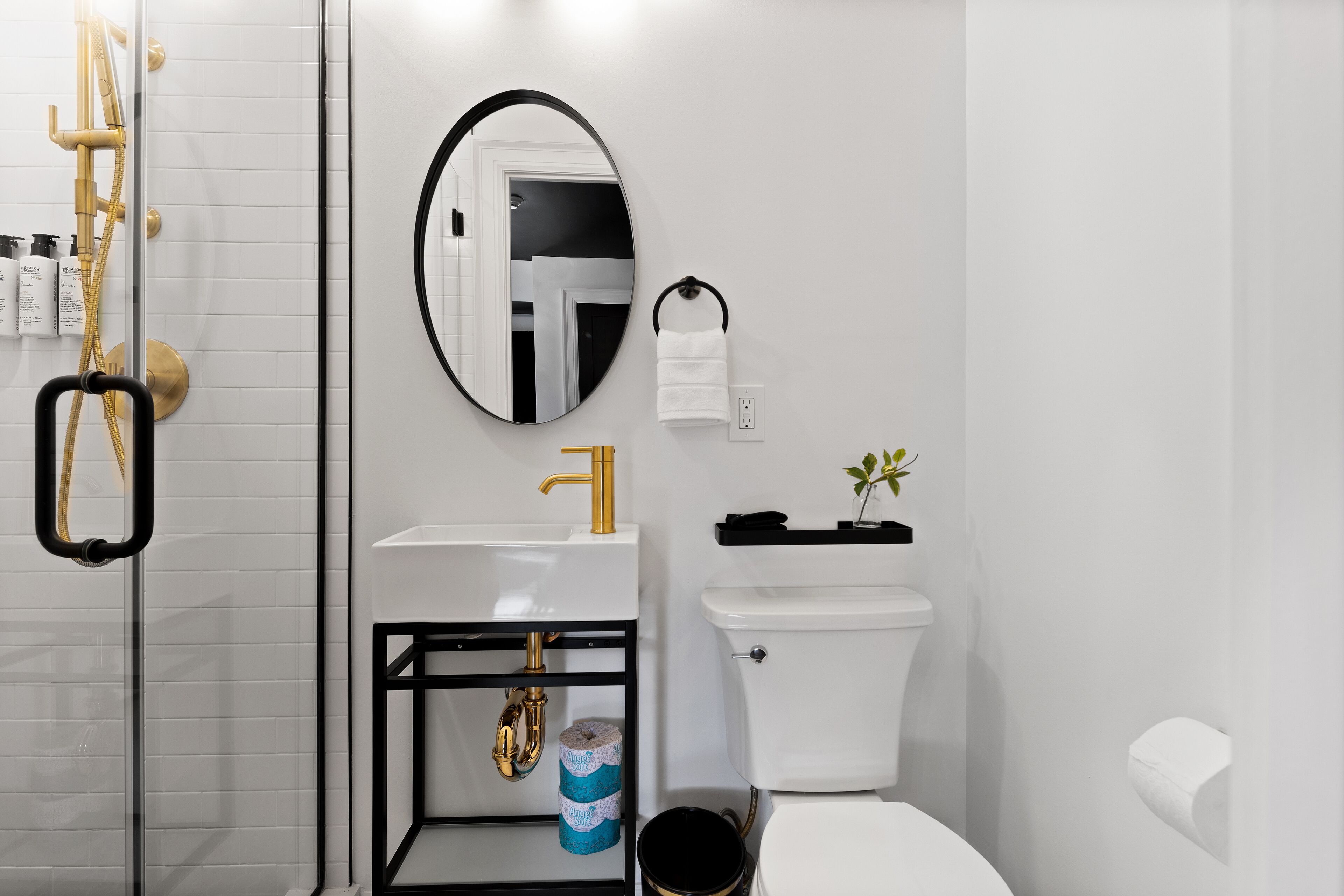 Un baño moderno con cabina de ducha de vidrio, un lavamanos blanco con detalles dorados, un inodoro y una pequeña estantería con artículos de aseo.