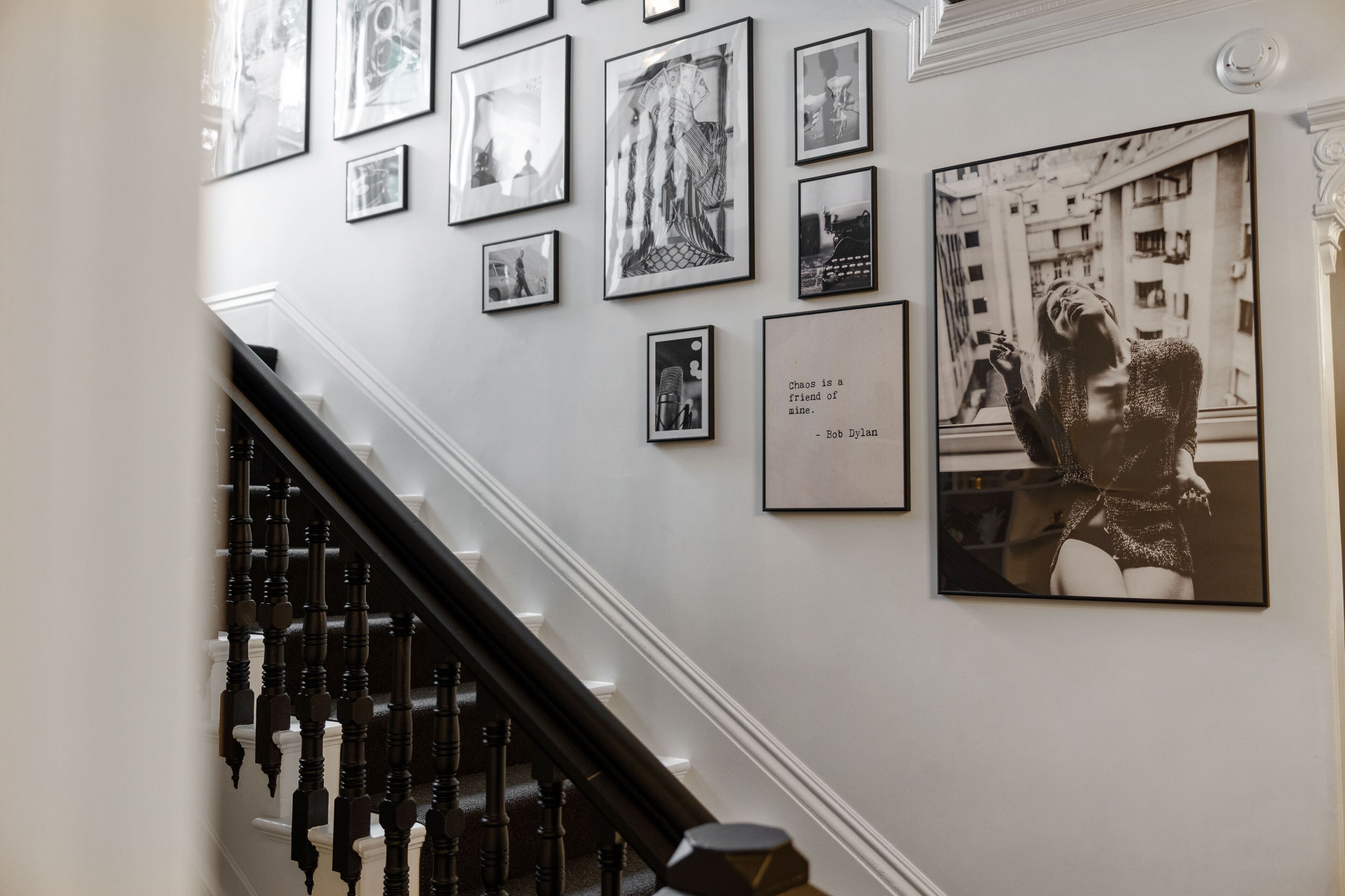 A staircase with a black handrail and white balusters, leading to a landing with a wall adorned with framed black and white photographs and a quote by Bob Dylan.