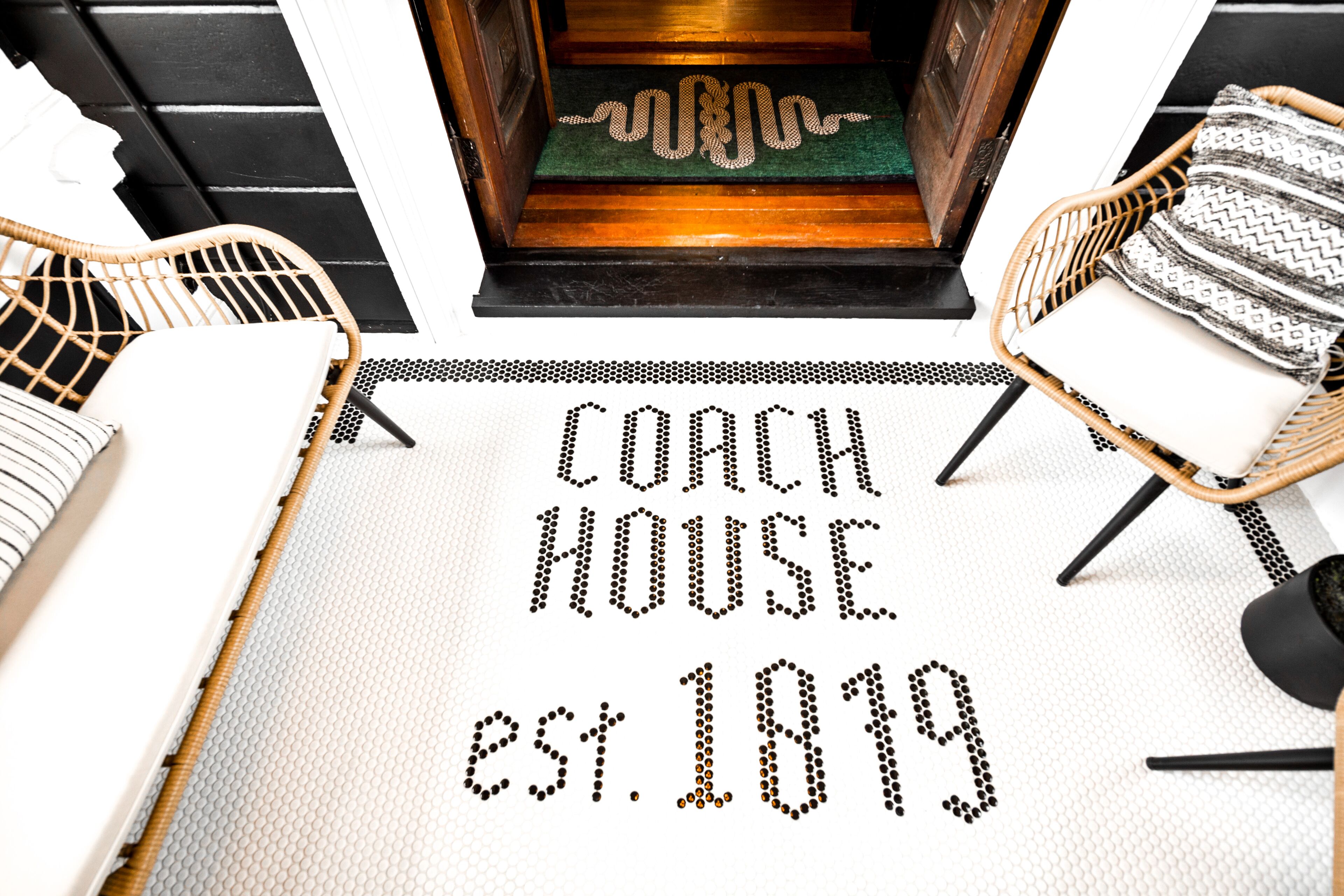 Um tapete com o texto "COACH HOUSE EST. 1819" em padrão de pontos, ladeado por cadeiras de vime com almofadas.