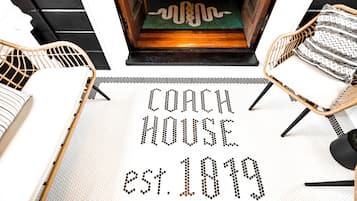 Um tapete com o texto "COACH HOUSE EST. 1819" em padrão de pontos, ladeado por cadeiras de vime com almofadas.