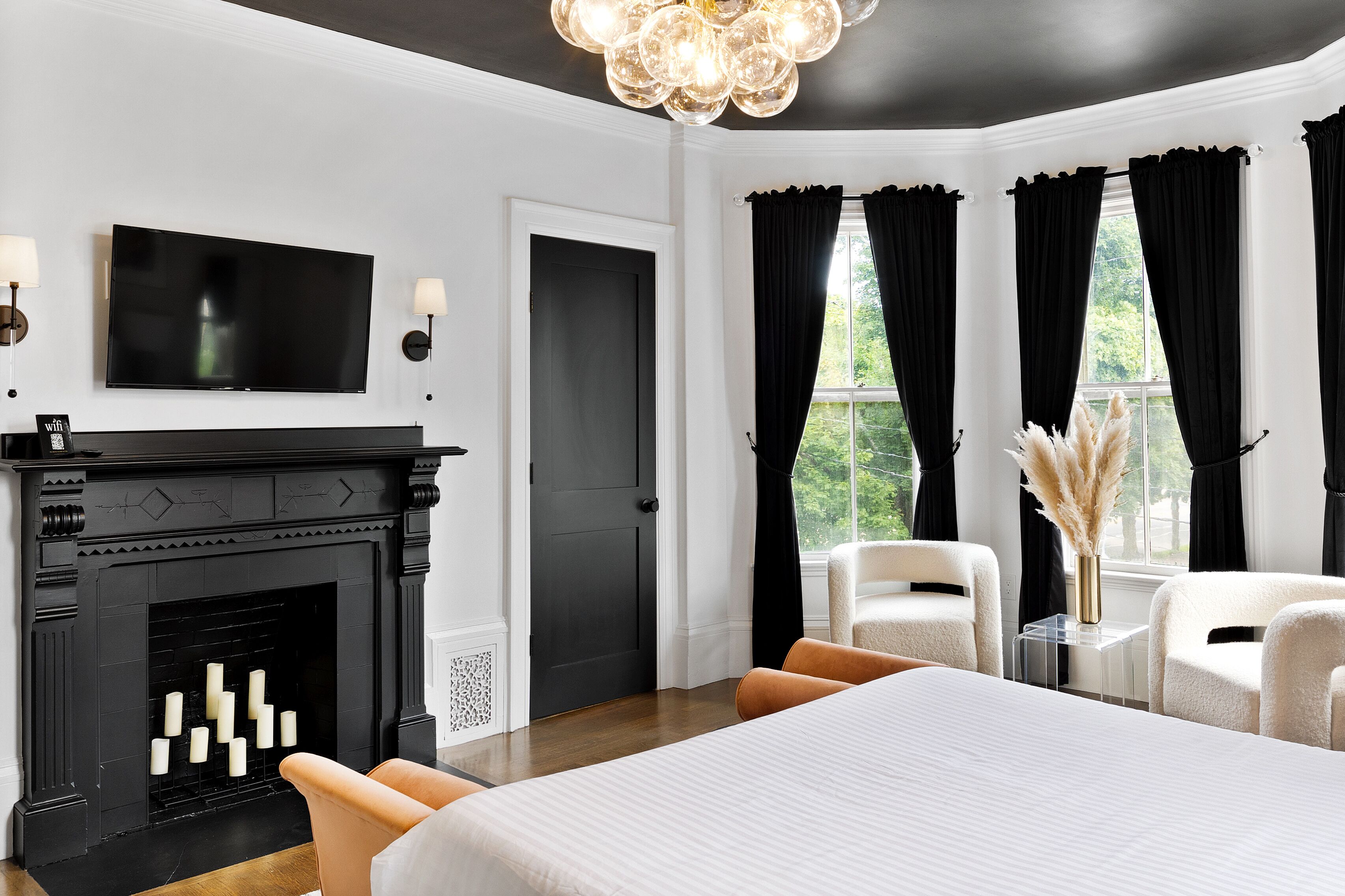 Una habitación de hotel moderna con una cama grande, una chimenea, dos sillones, un televisor y vistas a zonas verdes a través de las ventanas.