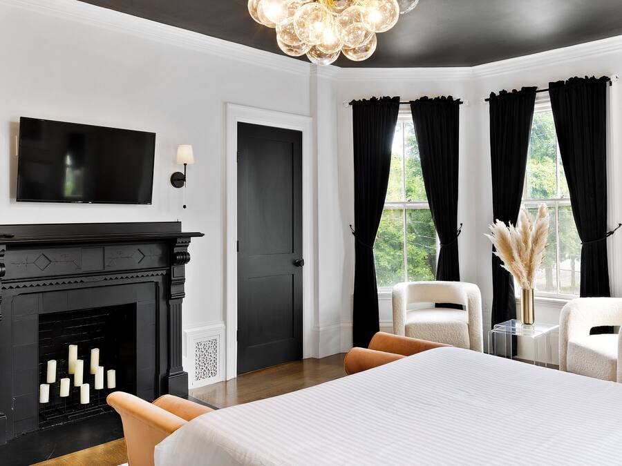 Una habitación de hotel moderna con una cama grande, una chimenea, dos sillones, un televisor y vistas a zonas verdes a través de las ventanas.