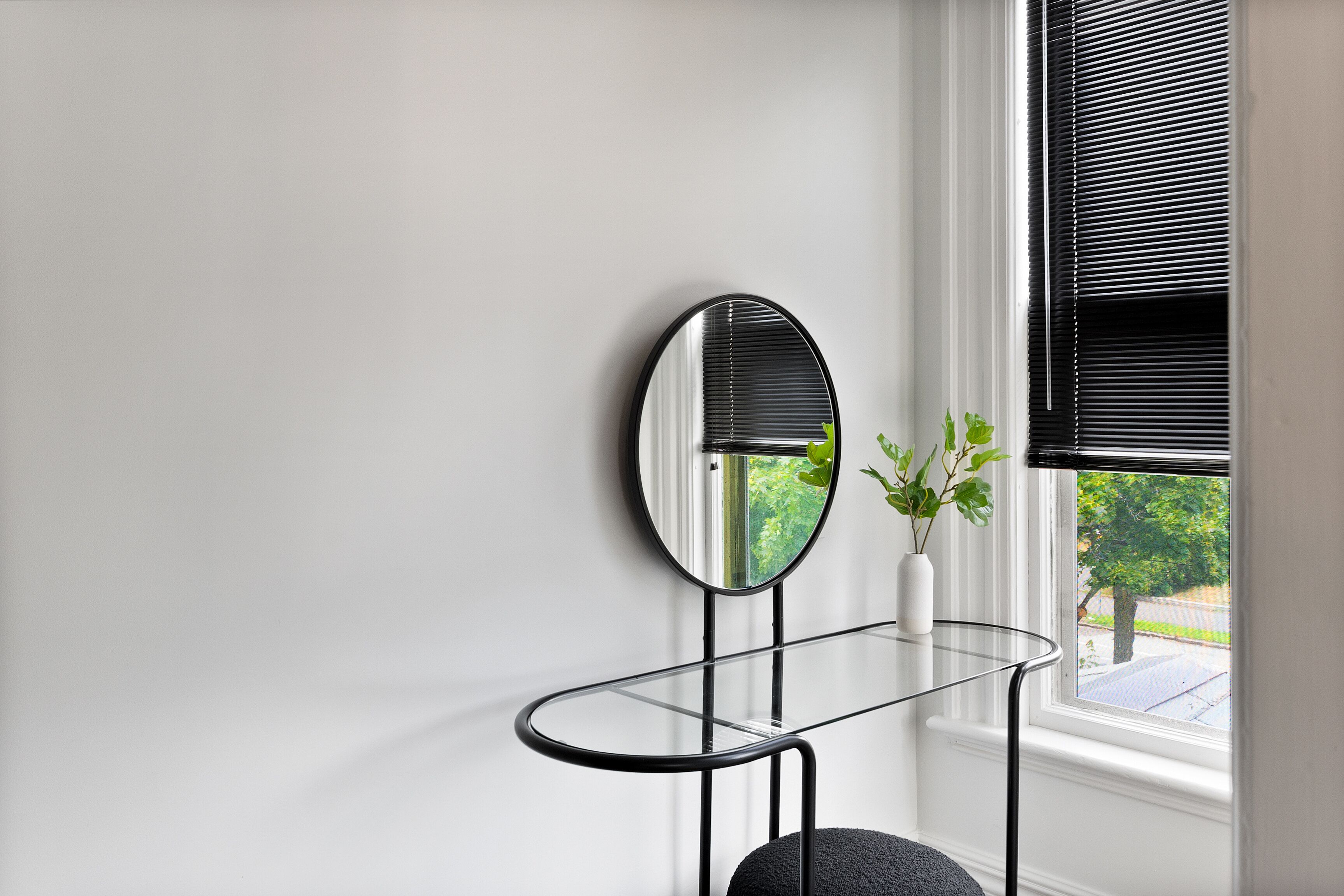 A corner with a round mirror on a glass table, a small potted plant, and a window with blinds.