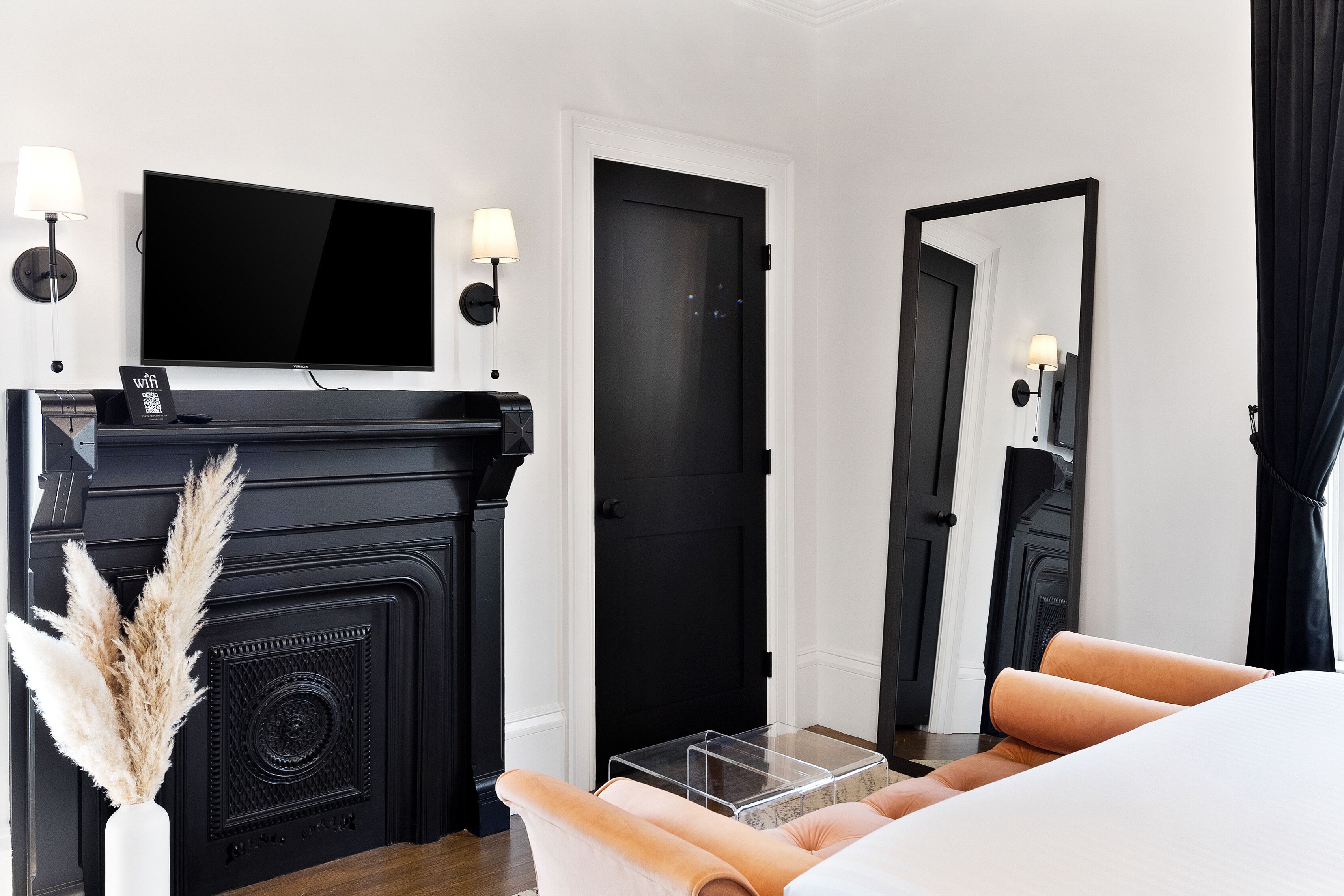 Una habitación de hotel moderna con una chimenea negra, un televisor de pantalla plana, un espejo grande y una cama con cubrecama blanco.