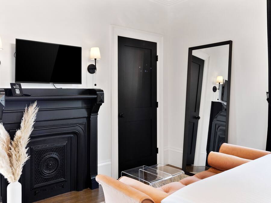 Una habitación de hotel moderna con una chimenea negra, un televisor de pantalla plana, un espejo grande y una cama con cubrecama blanco.