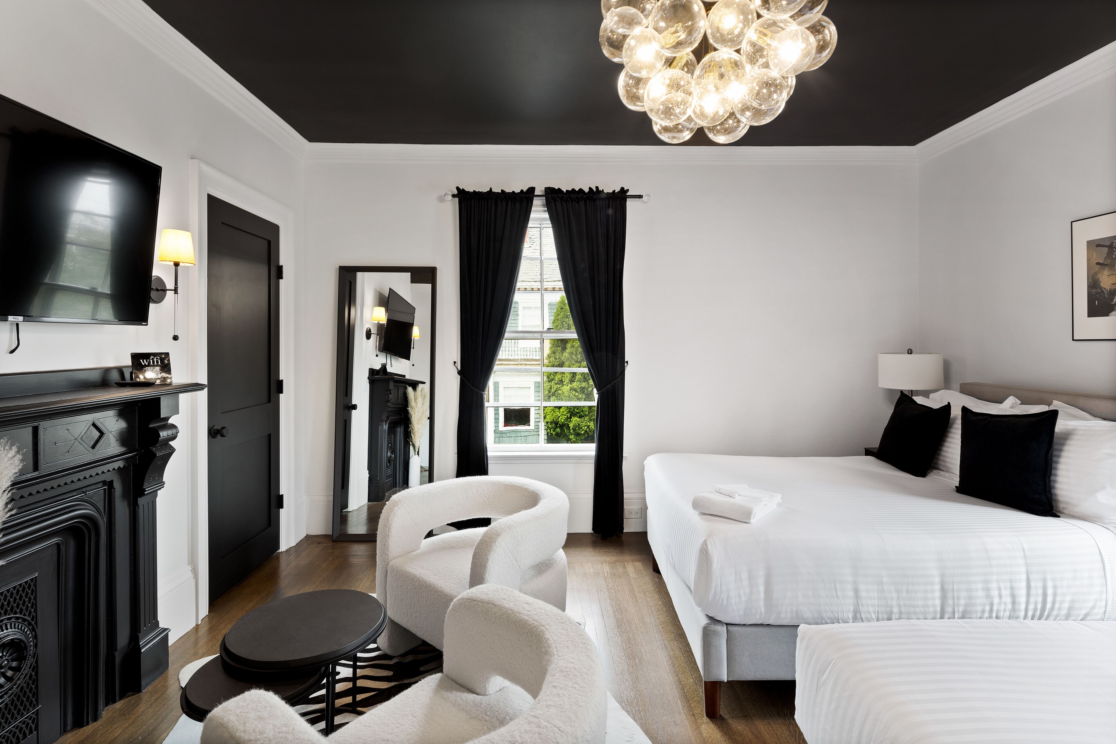 A modern hotel room with a black ceiling, a large flat-screen TV, a fireplace, a bed with white bedding, a sofa, and a view of the outdoors through a window with black curtains.
