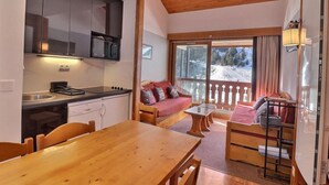 Eine kompakte Küche mit modernen Geräten, eine Essecke mit Holztisch und Stühlen sowie eine gemütliche Sitzecke mit Sofa und Blick auf eine verschneite Landschaft durch große Fenster.
