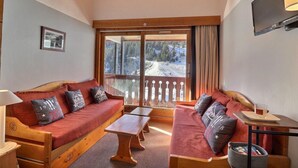 Ein gemütliches Zimmer mit Holzbalkendecke, einem großen Fenster mit Blick auf einen verschneiten Berg, einer Couch mit Kissen, einem kleinen Tisch und einem an der Wand befestigten Fernseher.