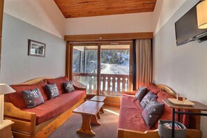 A cozy room with a wooden ceiling, a large window with a view of a snowy mountain, a sofa with cushions, a small table, and a TV mounted on the wall.