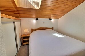 A bedroom with a wooden ceiling, a single bed, a nightstand with a lamp, and a small window.